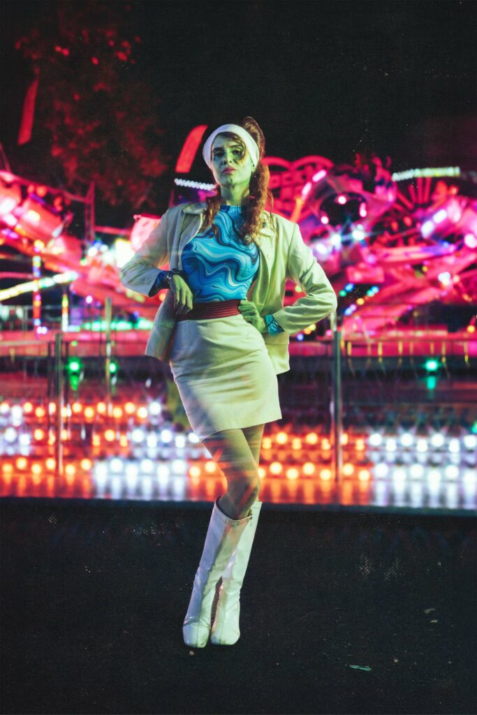 Portrait en-pied d'une femme qui prend la pose dans une fête foraine, vêtue d'un tailleur année 60, sa peau est verte, derrière elle, la lumière néon des attractions, shooting vintage