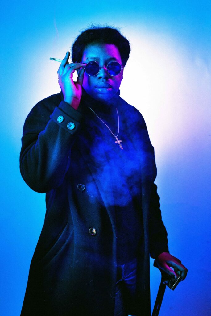 portrait gothique d’une femme sous lumières néon rose et bleu, lunettes noires et cigarette à la main