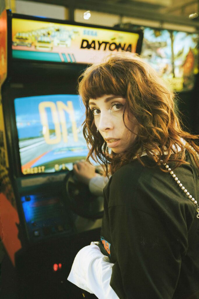 Portrait d'une femme rousse jouant à une borne d'arcade, tête tournée vers l'objectif, ambiance rétro.