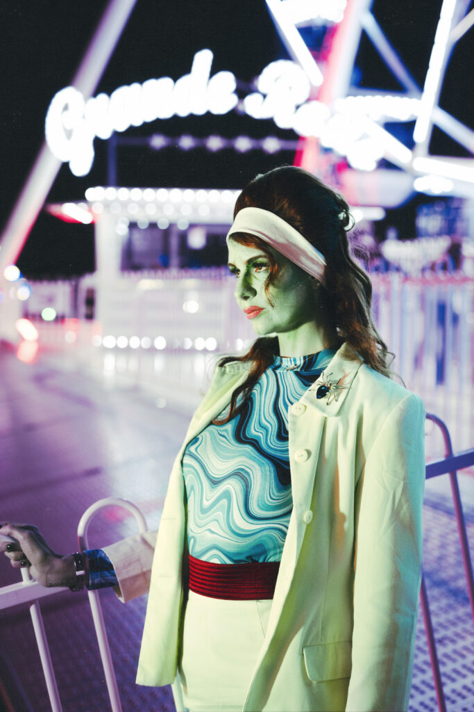 portrait d'une femme habillé années 60, la peau verte comme un alien de Venus, elle se tient dans une fête foraine.