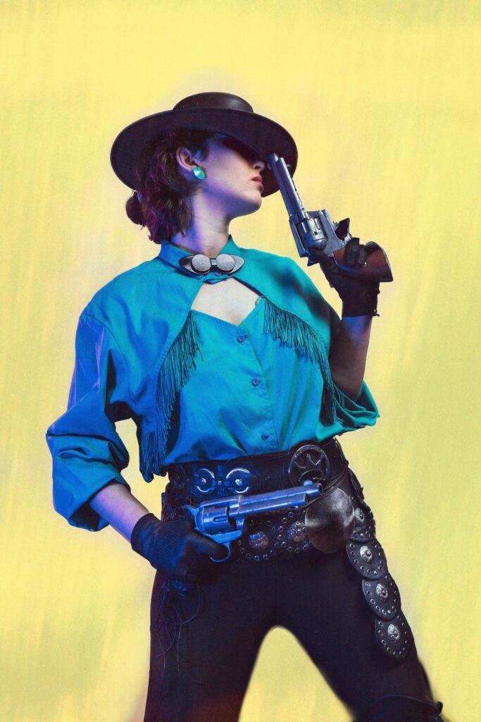 Portrait cinématographique d'une femme en habits de cow boy, pantalon de cuir, ceinture en métal, chemise bleu, elle porte un chapeau qu'elle relève avec un revolver