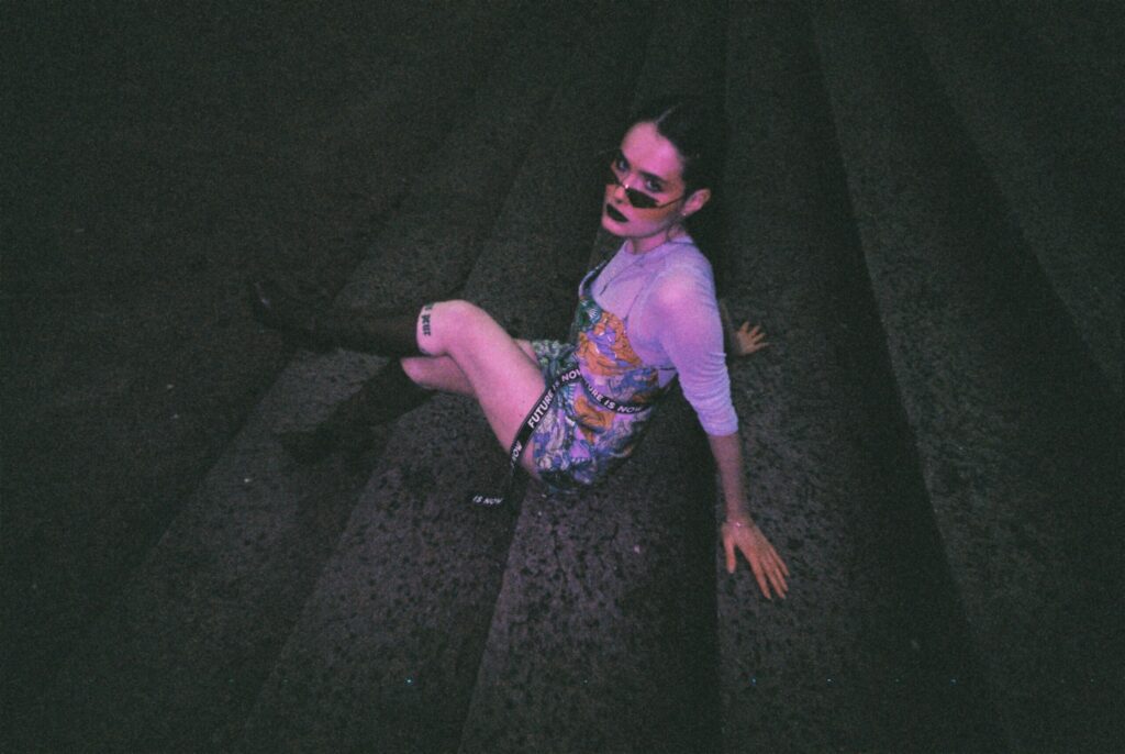 portrait d'une femme assise sur un escalier en pierre, en train de regarder l'objectif. Elle est éclairée en lumière violette.