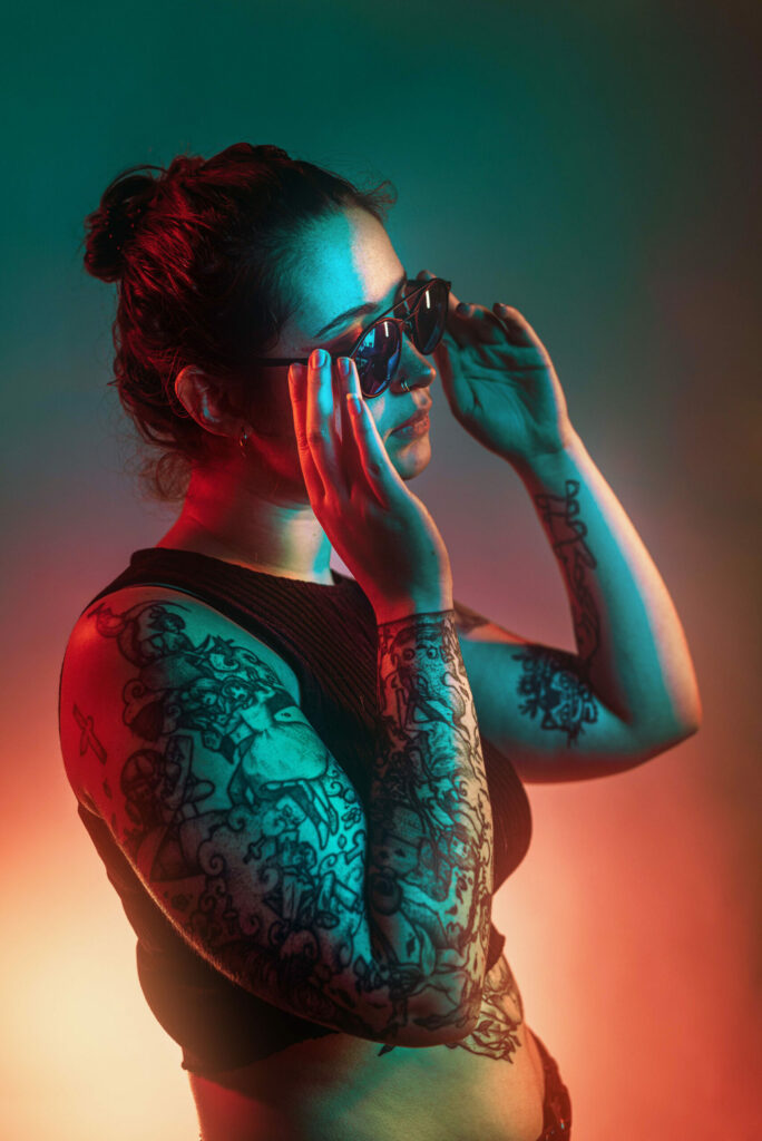 Portrait d’une jeune femme tatouée éclairée au néon, en train d’enfiler des lunettes de soleil, style cinématographique et mystérieux