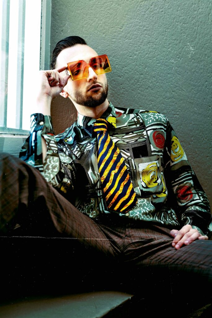 Portrait éditorial et vintage d’un homme au style très années 90, portant une chemise bariolée, une cravate rayée et des lunettes jaunes translucides.