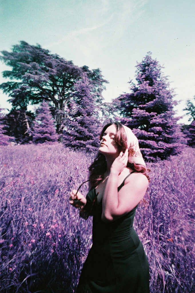 Portrait psychédélique d'une femme se recoiffant dans un champ aux sapins, herbe et arbres violets, style vintage pellicule années 70.
