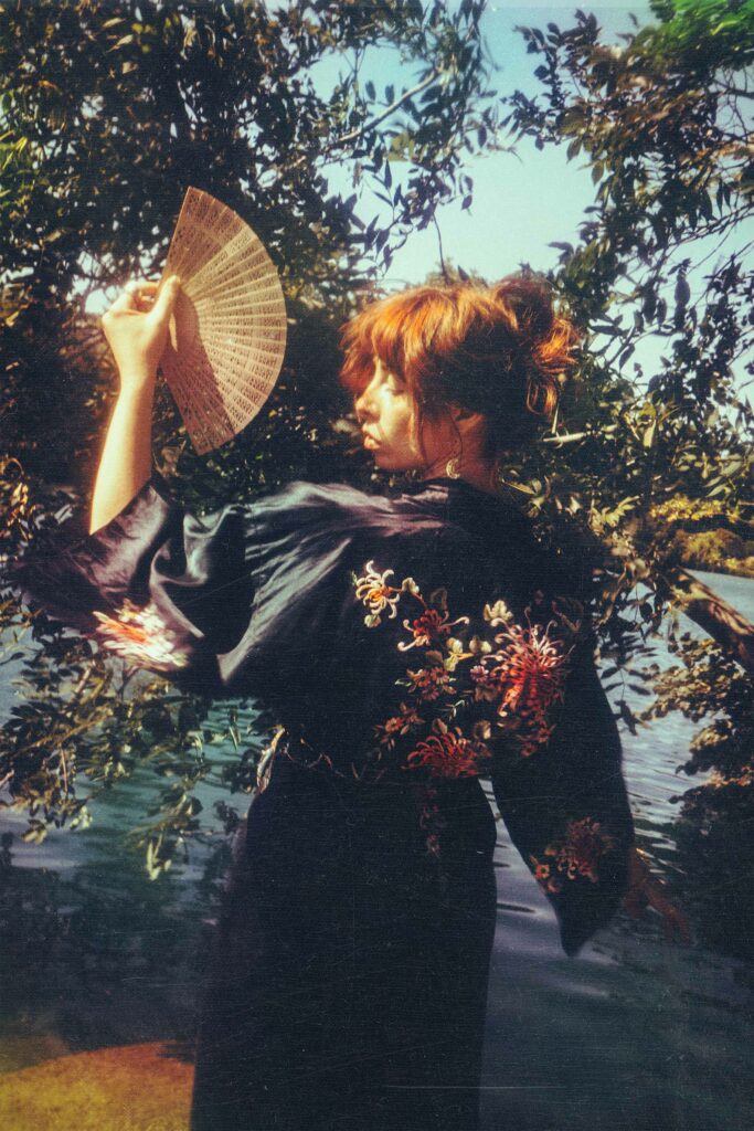 Portrait d'une femme de profil, portant un kimono noir à motifs floraux et tenant un éventail ouvert, ambiance estivale et ensoleillée, dans la nature