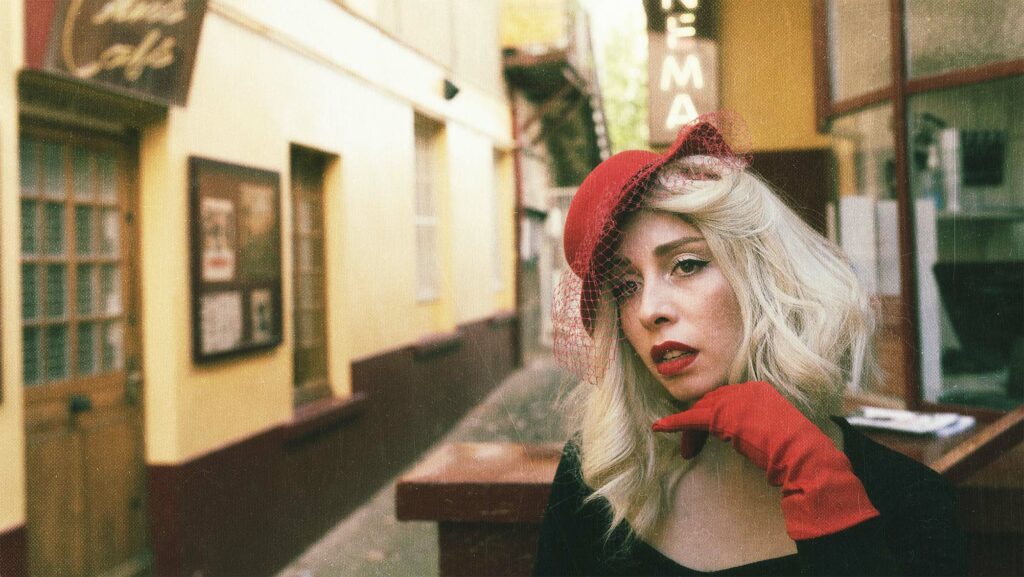 portrait d'une femme blonde portant chapeau rouge et gant rouge, elle attend devant un cinéma vintage
